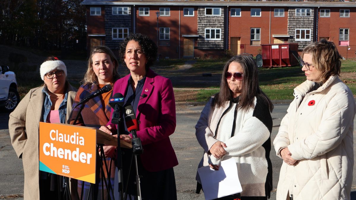 Five white women stand behind a podium that is set in a parking lot with abandoned and boarded up townhouses in the background. One of the women, who is wearing a fuchsia jacket over a navy dress is speaking into the microphone at the podium. An orange sign on the front of the podium says "Claudia Chender, NDP."